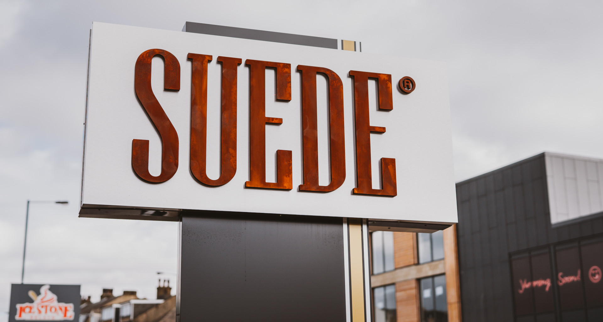 A signboard with the text ‘SUEDE’ in bold, brown letters against a cloudy sky, with other building signs visible in the background.