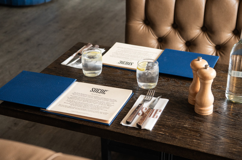 A set table at a restaurant with menus, glasses of water, cutlery wrapped in napkins, and a pepper grinder.